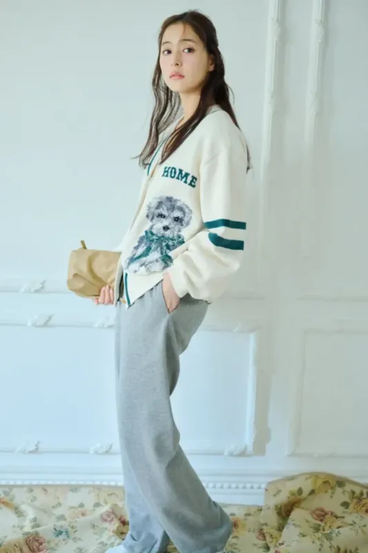 女性が犬のイラストが描かれたカーディガンとグレーのパンツを着て立っている写真です。