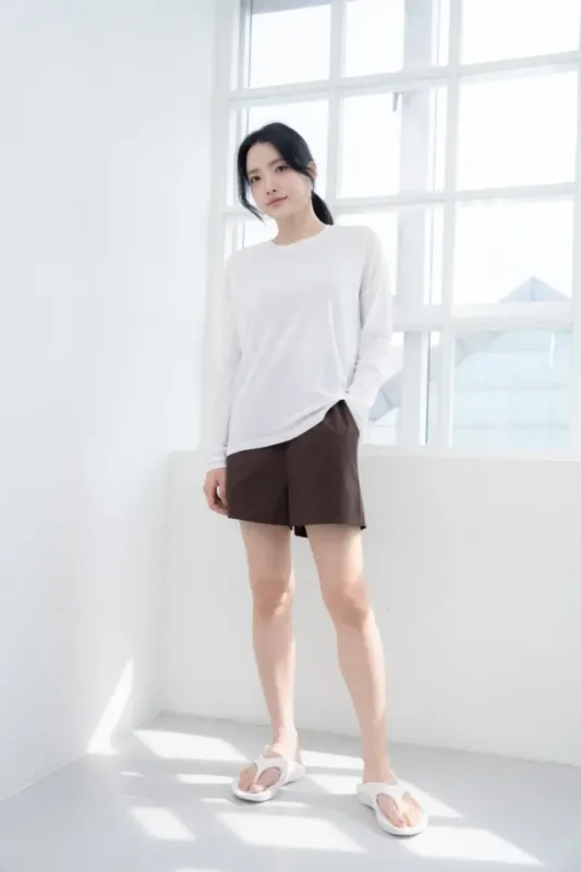 白い長袖シャツと茶色のショートパンツを着て、窓の前に立っている女性の写真です。彼女は白いサンダルを履いています。