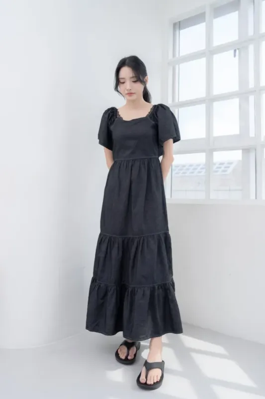女性が着ている黒いティアードワンピースの全身写真。女性は窓の前に立っており、彼女の顔は下を向いている。彼女は黒いサンダルを履いている。